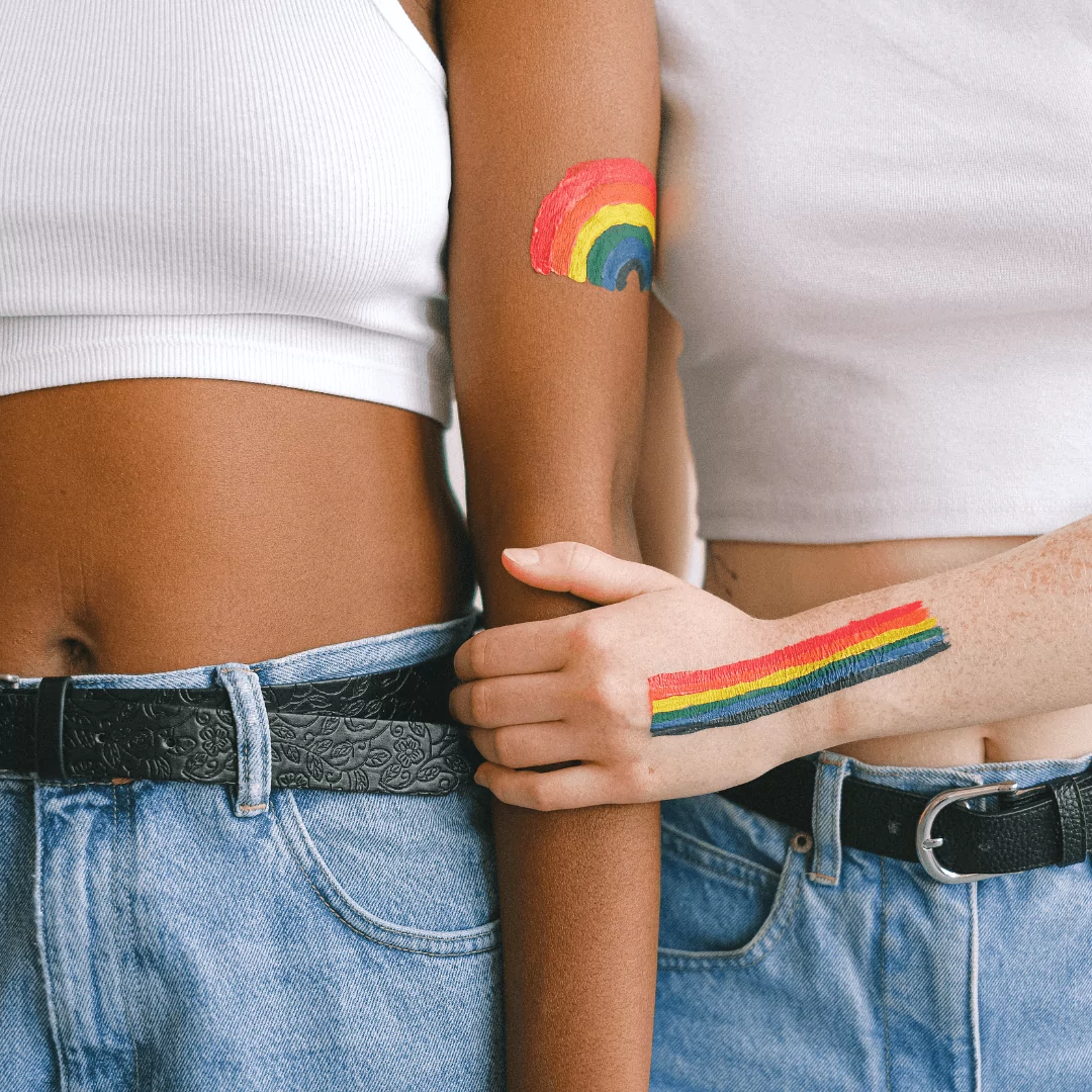 Zwei weiblich gelesene Personen mit aufgemalten Regenbögen halten sich am Arm